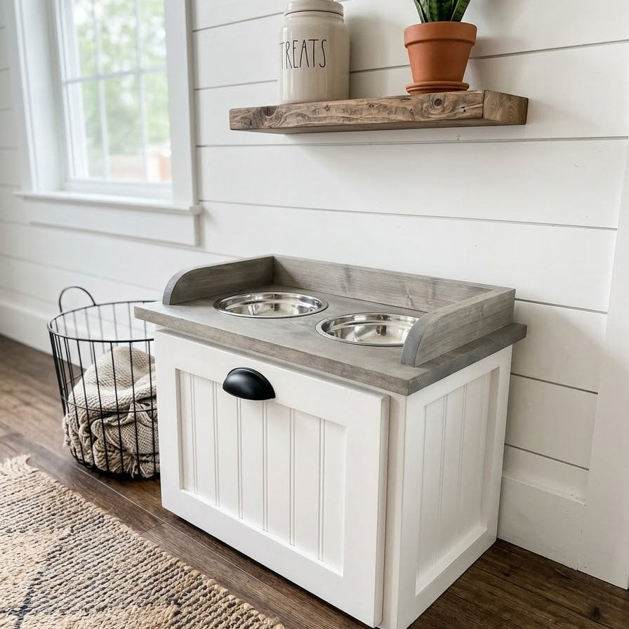 A white pet playhouse with a gray faux-wood top and two stainless steel bowls, set in a bright room with a window, wooden floor, and rustic shelves.