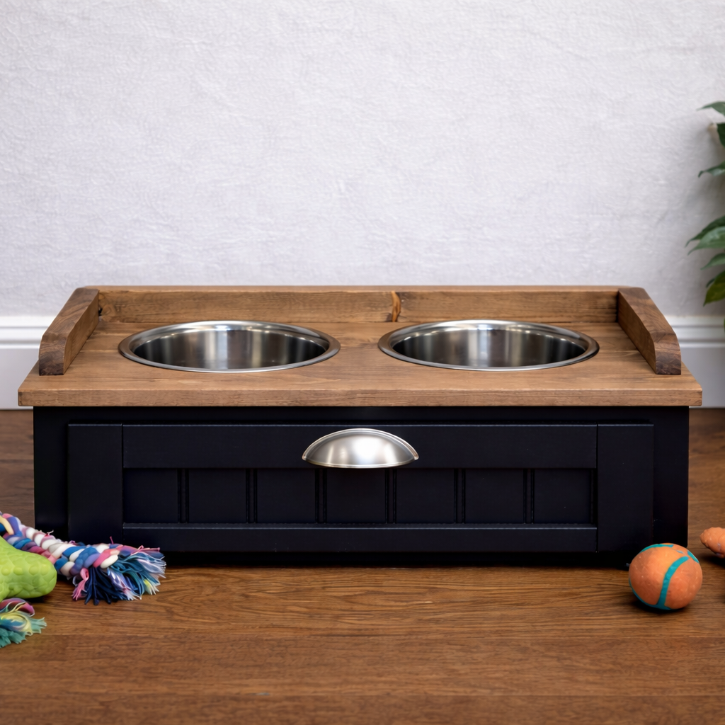 A black and wood elevated pet feeder and water bowl stand with two stainless steel bowls, shown on a wooden floor with dog toys and a potted plant in a room setting.